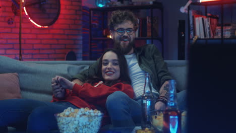 young happy boyfriend and girlfriend resting on the sofa at night, smiling and watching movie on tv