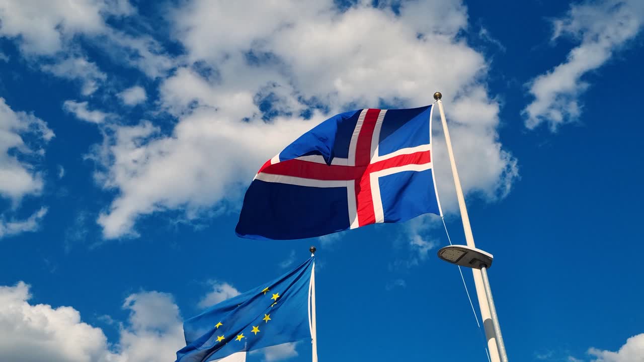 The Icelandic flag and the European Union flag sway gently in the wind beneath a blue sky scattered with white clouds