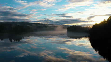 Aerial-shot-of-a-blank-river-running-through-a-pine-tree-forest-at-sunset-with-the-sky-reflecting-in-the-water