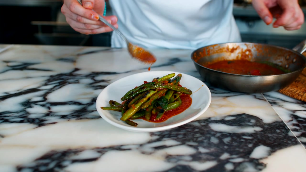Close-up de um chef profissional empratando uma refeição gourmet, adicionando cuidadosamente um molho vermelho ou guarnição de uma panela quente para um prato de cerâmica branco em um ambiente de cozinha de restaurante