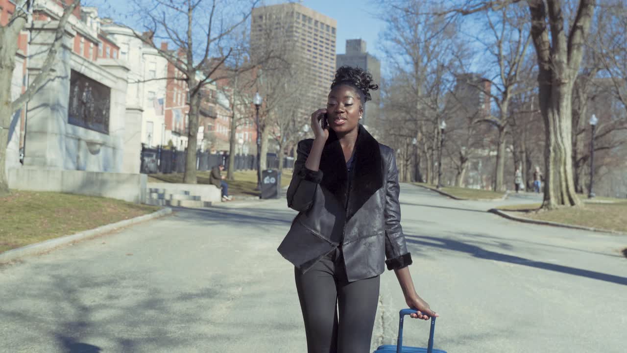 Fashionable Young African American Business Woman Walking Through Park With Luggage While Talking On Phone Traveling On A Sunny Day