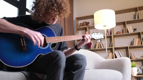 a curly-haired guy is sitting on a sofa and playing an acoustic guitar, he has a headache. migraine and headache in a young man