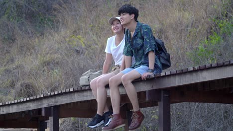 4k relaxed young asian couple man and woman hiker sitting and enjoying together on wooden footpath bridge at the mountain. relationship and outdoors lifestyle in summer vacation concept.