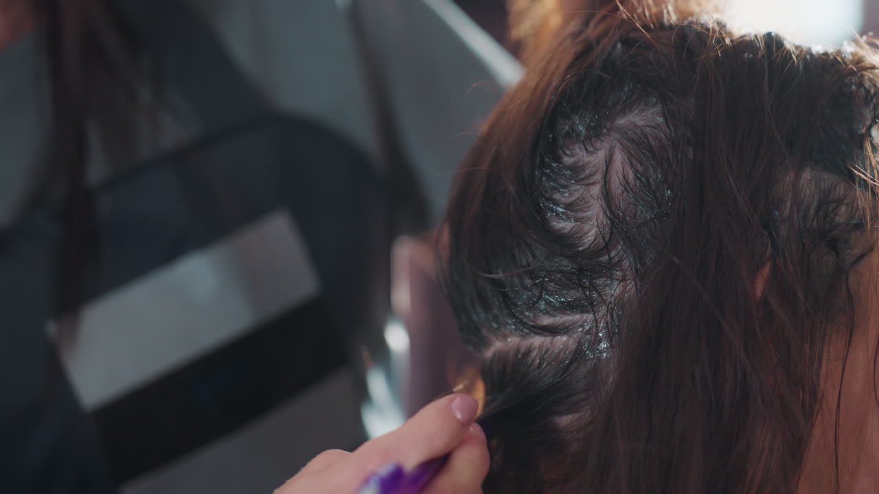 코카서스인 컬러리스트가 보라색 어플리케이터 브러시를 사용하여 가르마가 있는 뿌리에 염색약을 펴 바르고, 세심한 터치와 섹션 컨트롤을 하고 있어요. 햇빛이 질감 있는 머리카락을 비추고, 현대적인 살롱 스테이션이 배경에 보입니다.