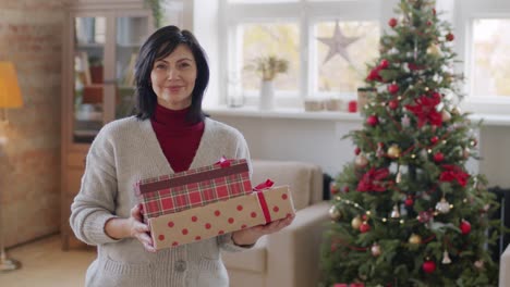 brunette woman in casual clothes looks at camera and smiles while holding christmas gifts