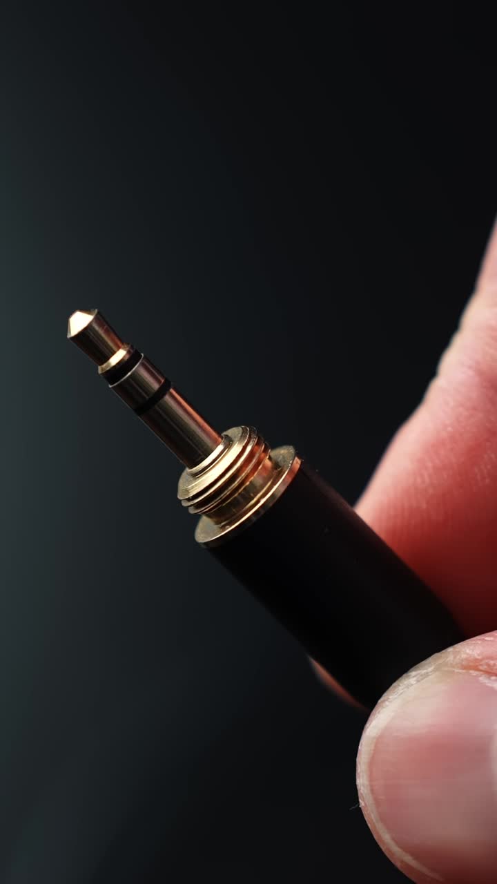 Warm macro close-up of a 3.5mm headphone jack held between fingers on a dark background, sharp metal detail with a calm, minimal tech vibe