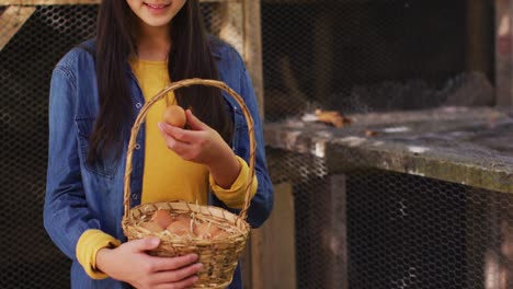 smiling asian girl with basket of eggs standing by hen house in garden