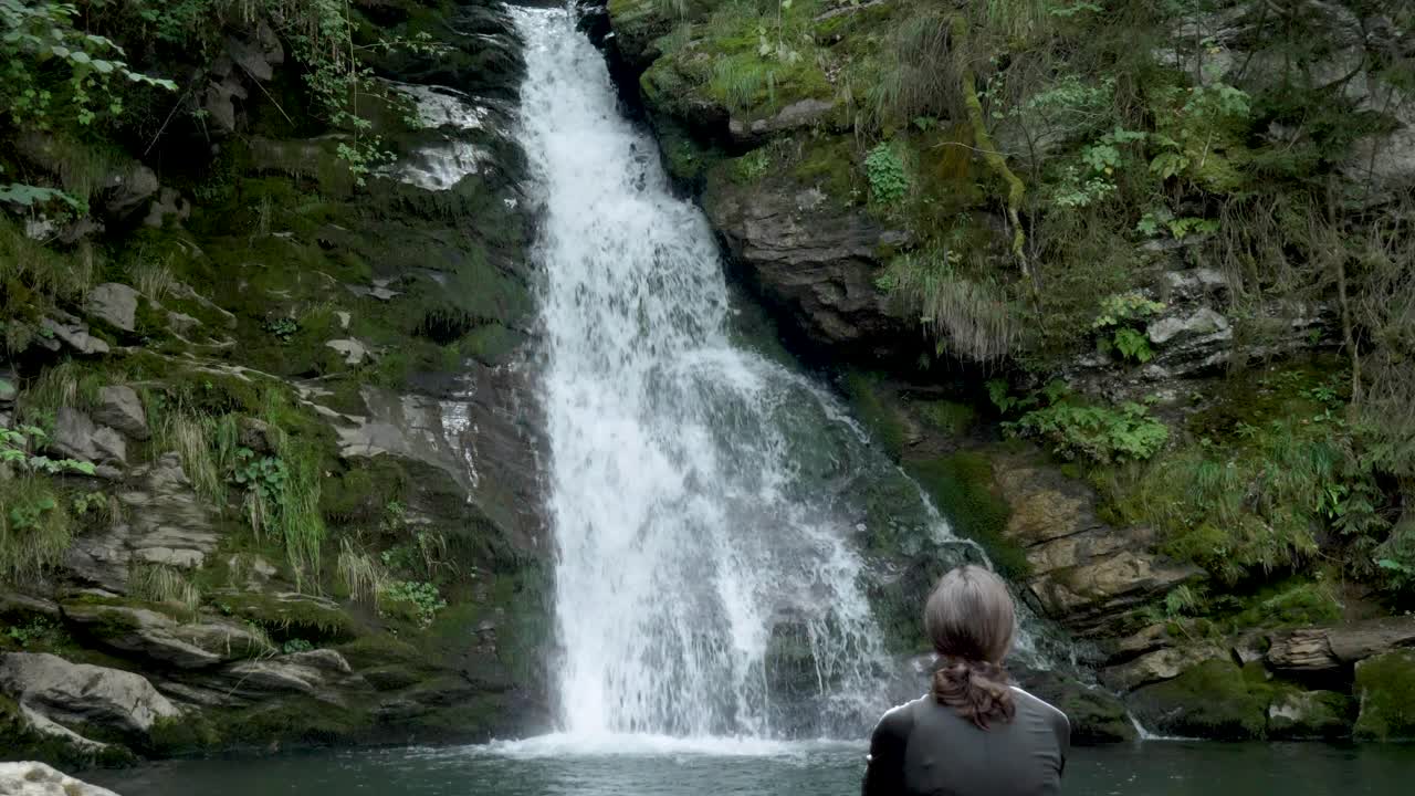 una mujer solitaria se sienta en contemplación frente a una cascada de bosque alpino y observa el flujo de agua por la roca hacia el pequeño lago alpino y arroyo