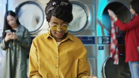 young pretty and stylish girl in yellow glasses standing in laundry service room and flipping pages of fashion journal