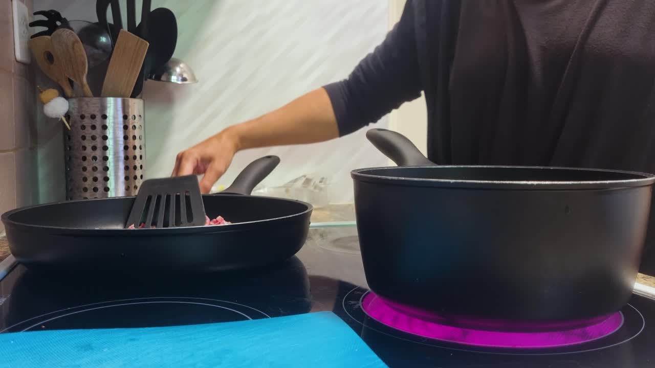 Close-up of a woman’s hands in a cozy Swiss kitchen as she fries bacon in a black pan, stirring with a plastic utensil while water boils in a second pot for a homemade meal