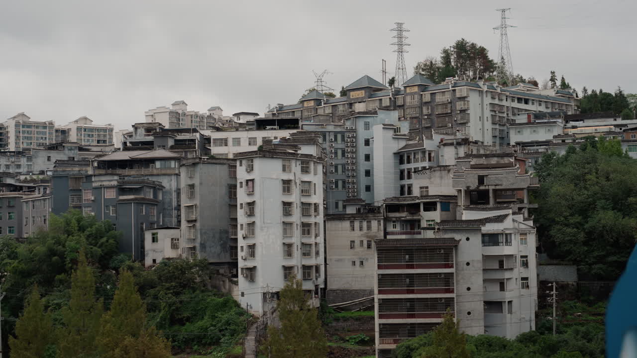 Skyline urbano collinare sotto un cielo coperto. Prospettiva da drone di densi blocchi residenziali con facciate in cemento invecchiato, antenne raggruppate. Alberi sparsi in primo piano, palette di colori tenui, atmosfera pesante.