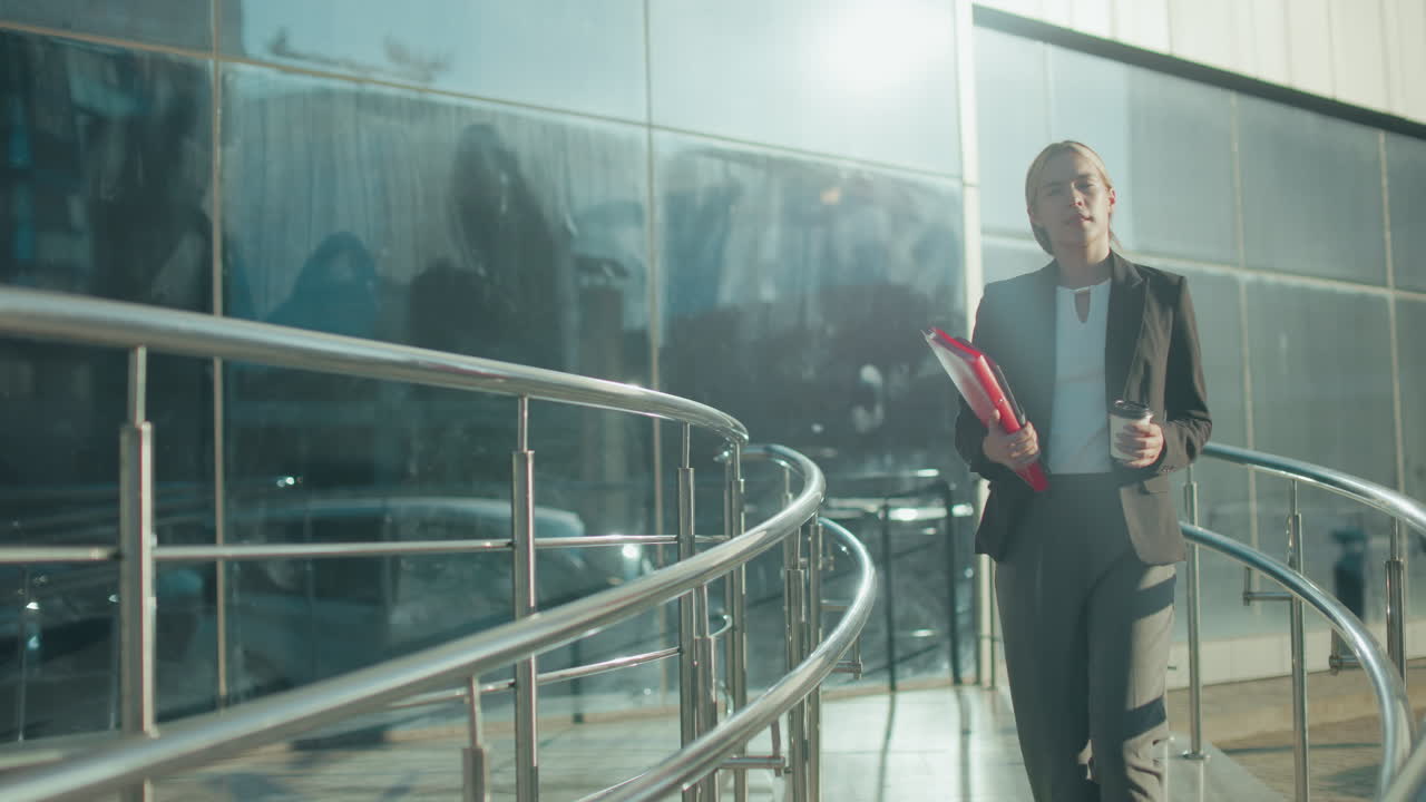 Young lady holding folder and tea cup walking along path with iron railing in sunlight near glass building reflecting ambient cityscape while dressed in formal outfit in bright urban setting