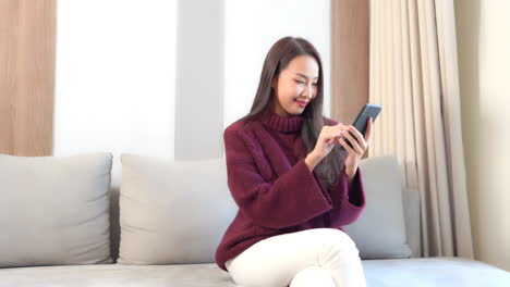 young pretty asian woman texting and chatting on smartphone at living room sofa