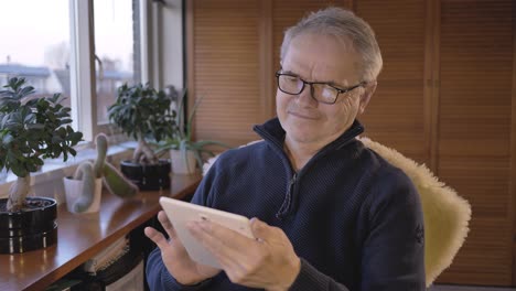 Middle-aged-caucasian-man-looking-through-photos-on-a-tablet-and-smiling-in-a-hobby-room-near-a-window-with-plants