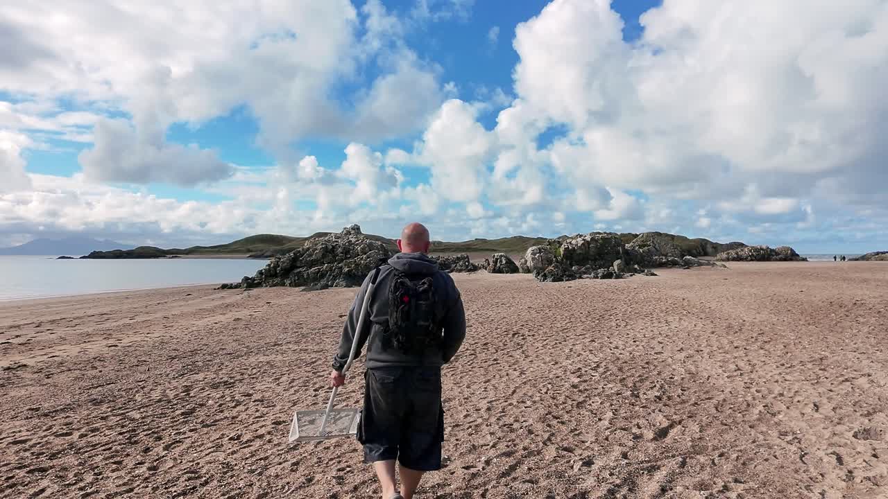 Bald male carrying sand scoop walking dog on sunny Welsh beach active lifestyle exploring