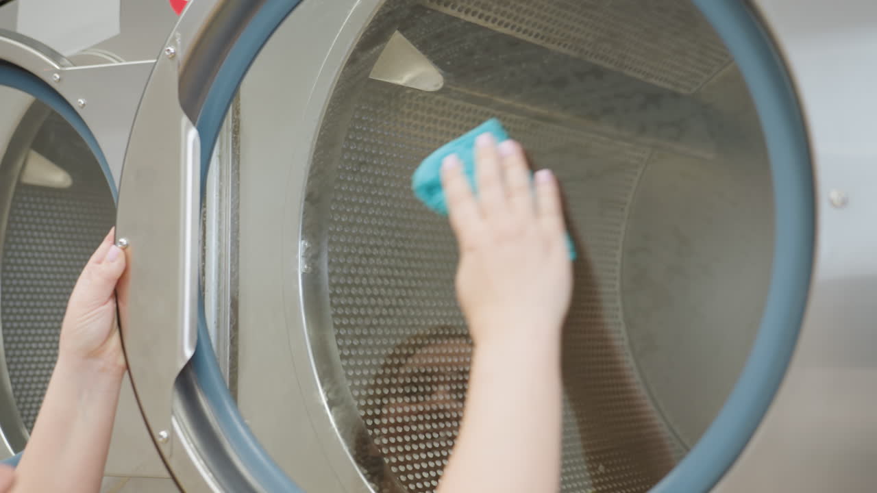 Laundry owner in yellow shirt cleans transparent surface of industrial washing machine with blue towel, reaching up to wipe glass door, reflection faintly visible on shiny metal surface