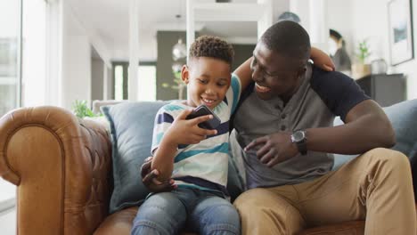 video of happy african american father and son sitting on sofa and using smartphone