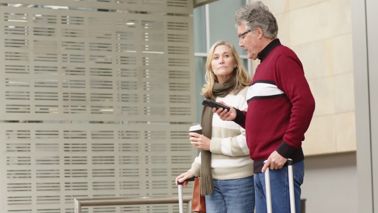 Senior couple heading terminal entry pulling luggage checking route and sipping coffee, copy space