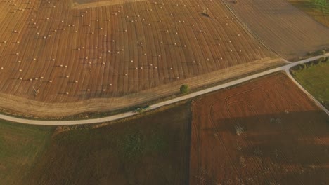 Tractor-With-Trailer-Drives-Through-A-Rural-Gravel-Road-Leading-Through-Agricultural-Fields-On-A-Summer-Evening