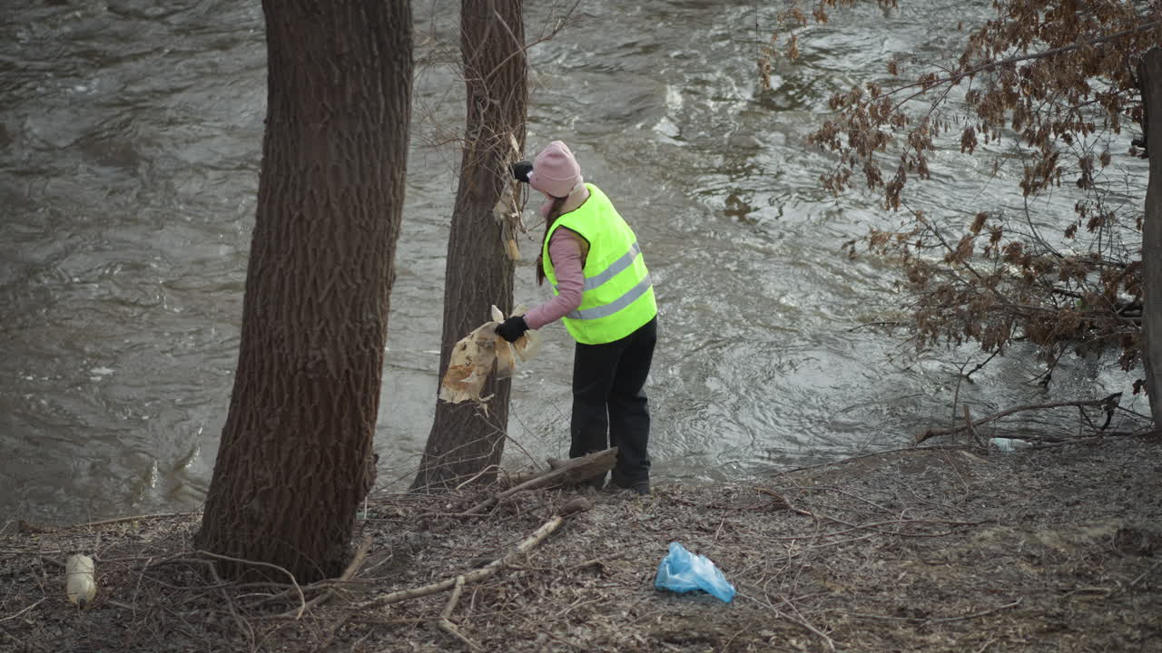 Woman in high visibility vest and pink beanie removes dirty plastic sheet tangled on tree trunk along fast-flowing river during environmental cleanup effort, holding debris in gloved hands