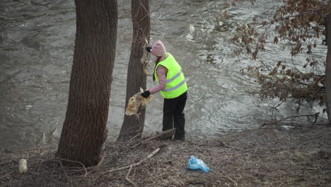 woman in high visibility vest and pink beanie removes dirty plastic sheet tangled on tree trunk along fast-flowing river during environmental cleanup effort, holding debris in gloved hands