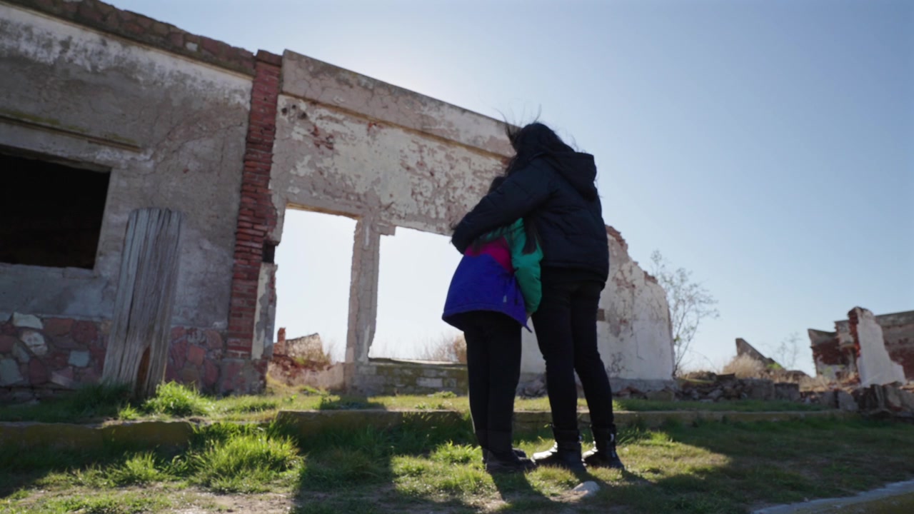 Inquadratura statica di madre e figlia che si abbracciano dietro un muro di cemento tra le rovine abbandonate di Villa Epecuén prima del tramonto.