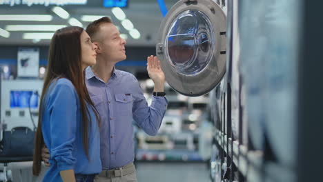 young man and woman in appliances store choose to buy washing machine for home. open the door looking into the drum, compare the design and characteristics of the devices.