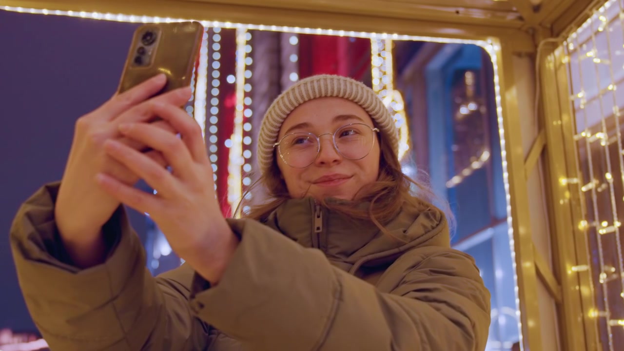 Ung kvinne tar selfie med telefon inne i opplyst julebod, scroller meldinger og ler, iført briller og vinterkåpe, varm glød og ekte sosial forbindelse fanget om natten.