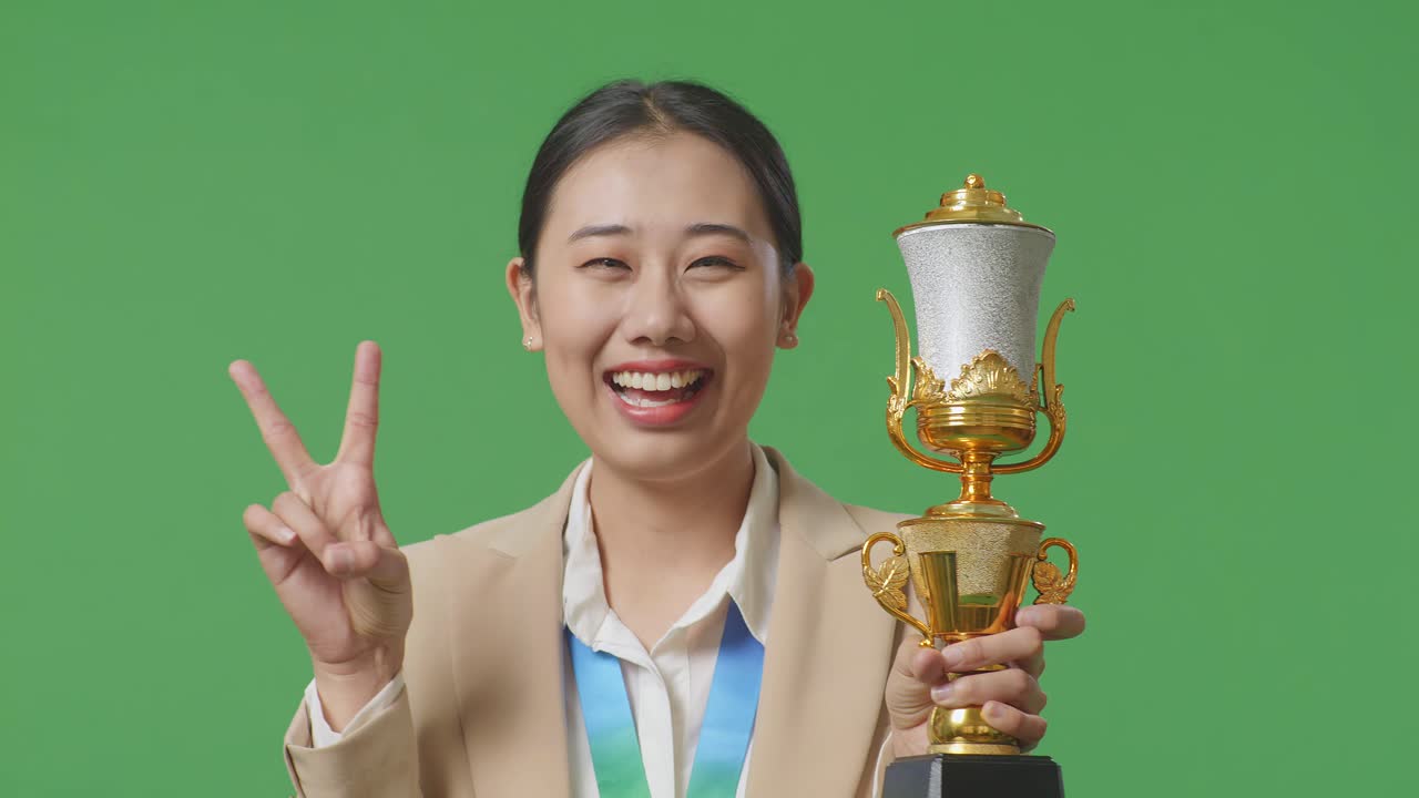 금메달과 트로피를 입은 슈트를 입은 아시아 사업가 여성이 평화의 몸짓을 보여주고 스튜디오의 녹색 스크린 배경에서 첫 번째 우승자로 카메라에 미소 짓고 있습니다.
