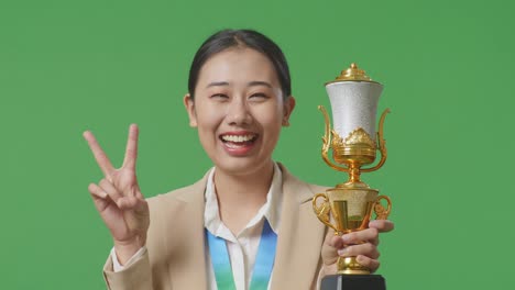 close up of asian business woman in a suit with a gold medal and trophy showing peace gesture and smiling to camera as the first winner on green screen background in the studio