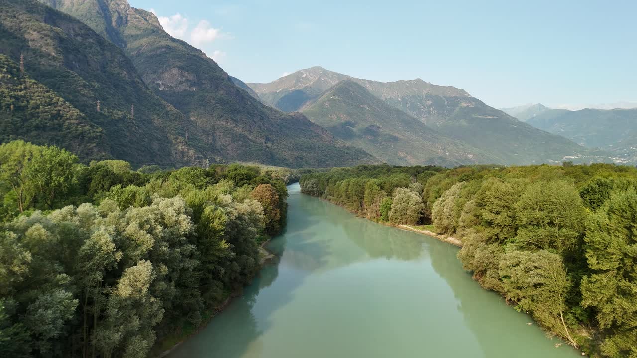 A fast drone flyover of the Toce River, framed by trees, with mountains and sky in the background, showcasing the stunning landscape of Ansola, Italy.