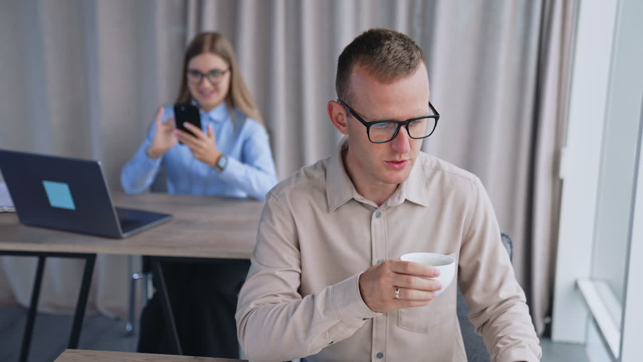 Young people working in the modern office. Man with cup of coffee works at laptop and woman using a phone at backdrop in blur.