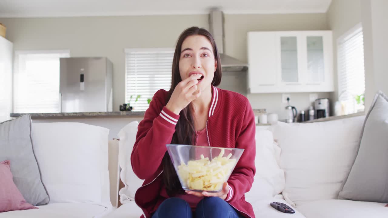 Donna caucasica felice seduta sul divano, che mangia snack e guarda la TV in salotto.