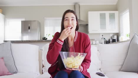 Glückliche-Kaukasische-Frau,-Die-Auf-Der-Couch-Sitzt,-Snacks-Isst-Und-Im-Wohnzimmer-Fernsieht