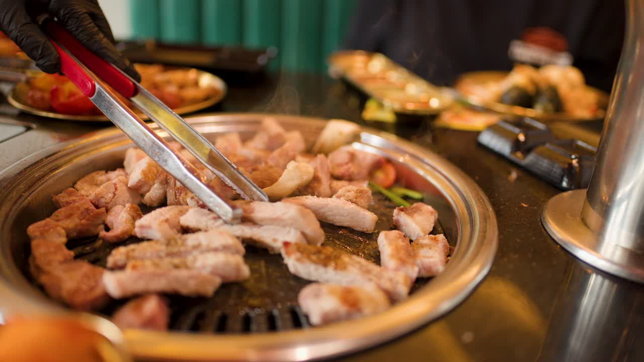 Person cuts and grills pork pieces on tabletop grill, steam rising, warm indoor lighting, close-up