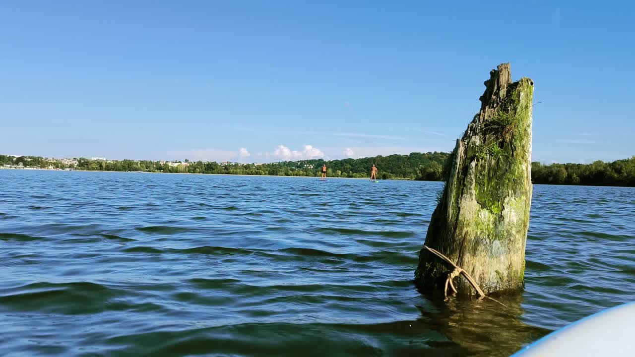 A steady lake view shows two paddleboarders moving across quiet water on a clear sunny day, with gentle ripples and open sky creating a relaxed natural scene as one paddles without clothing