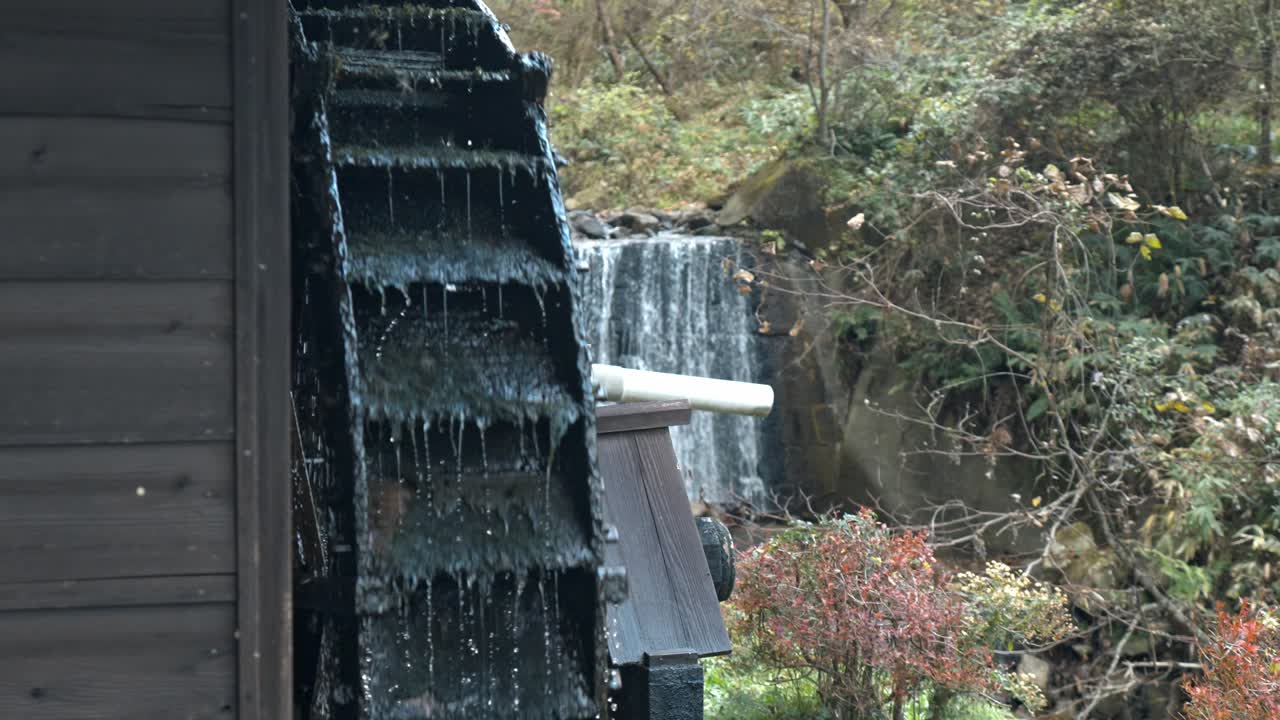 A serene view of a traditional water mill along the Nakasendo Trail in Japan, framed by a gentle waterfall in the background.