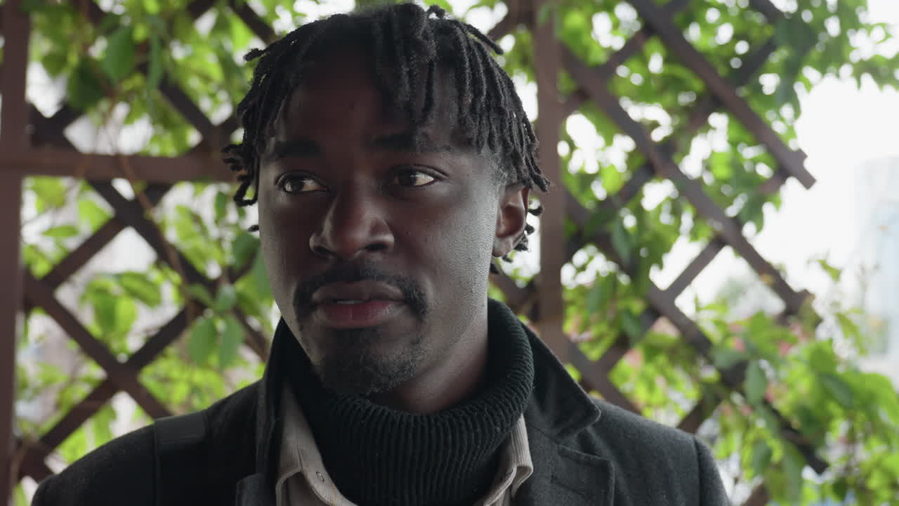 Camera view shows dreadlocks man backing crisscross iron grille, x pattern filling frame, head turning left then right, tensed yet curious mood, metal lattice forming bold graphic texture