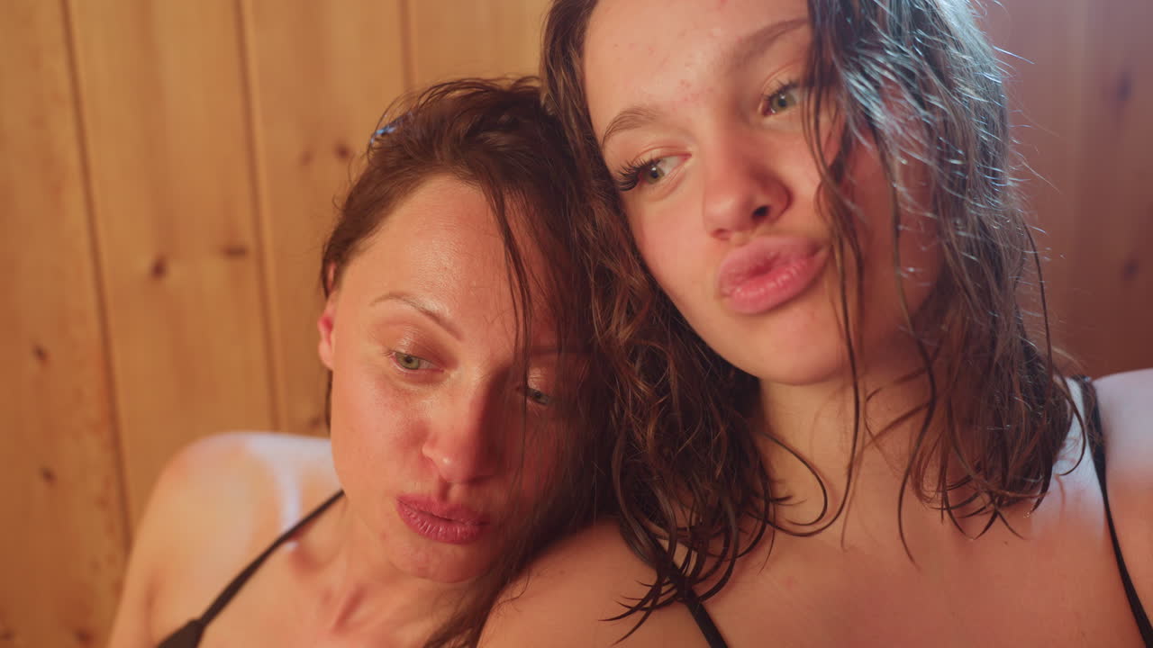Close up of woman and friend taking selfie against warm wood wall interior, soft back light highlighting wet hair and smiling faces, capturing joyful bond in intimate indoor vlog moment