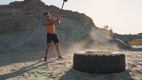 muscle athlete strongman man hits a hammer on a huge wheel in the sandy mountains in slow motion. strength and endurance training for wrestlers