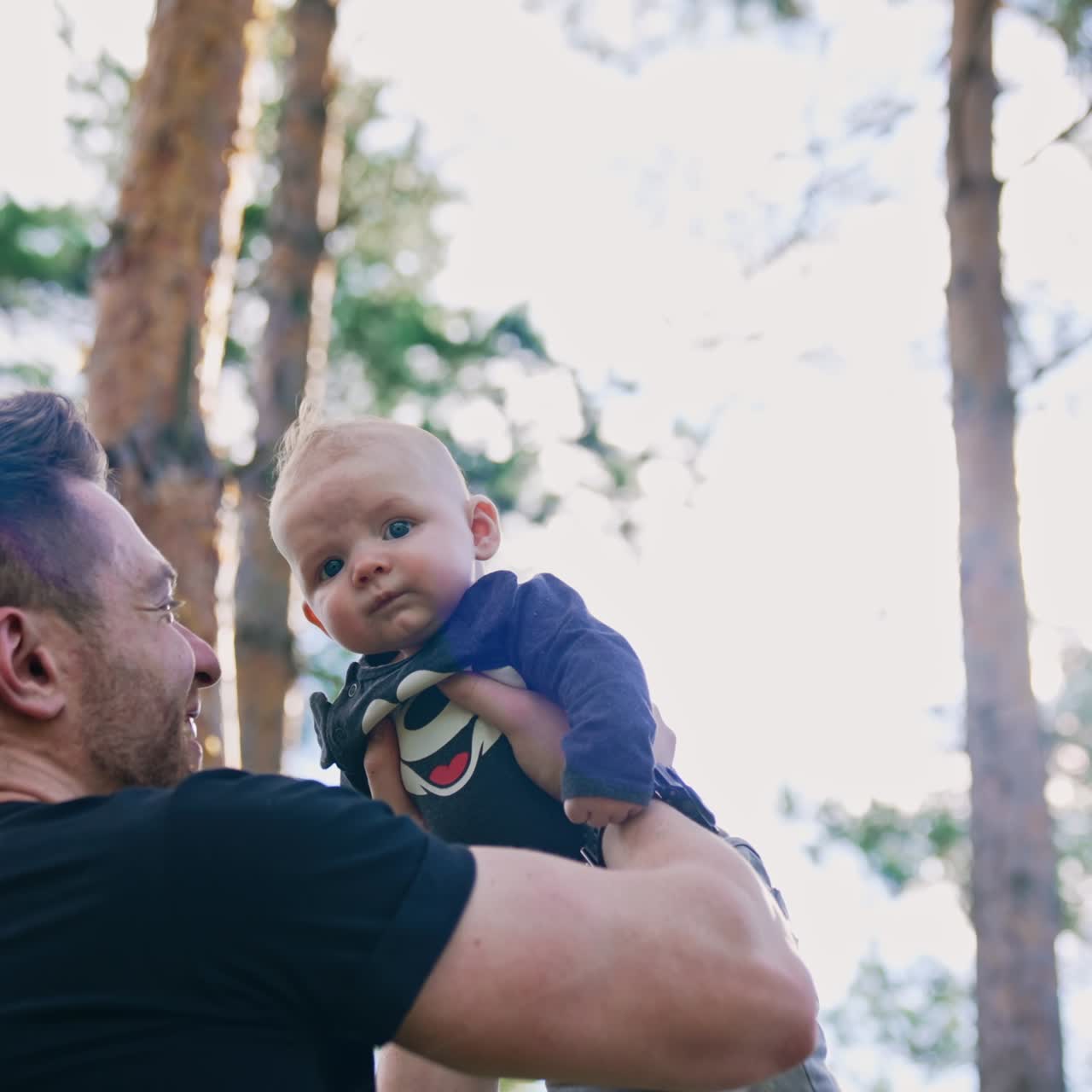 Bearded Caucasian man holding a cute newborn boy over the head. Happy dad looks with adoration on his baby. Low angle view. Pine trees at backdrop
