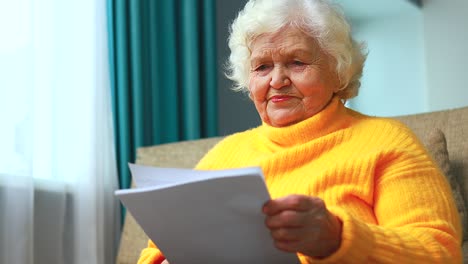 shoked amazed old woman white gray-haired sitting on the sofa in living room with bills