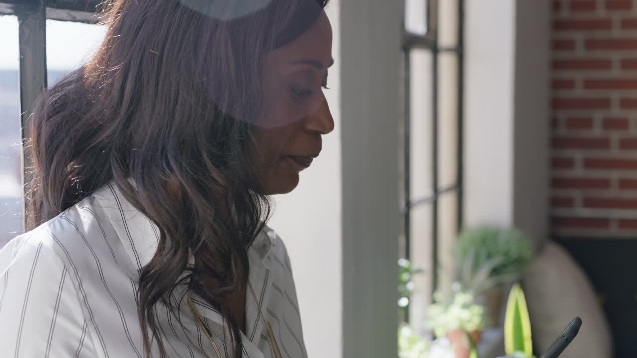 retrato de una mujer afroamericana feliz usando un teléfono inteligente bebiendo café en casa navegando por los mensajes de las redes sociales disfrutando de una mañana relajada mirando por la ventana enviando mensajes de texto en el teléfono móvil