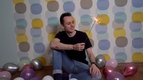 Young-Man-Sitting-with-a-Sparkler-Against-Colorful-Background,-Surrounded-by-Balloons-on-the-Ground