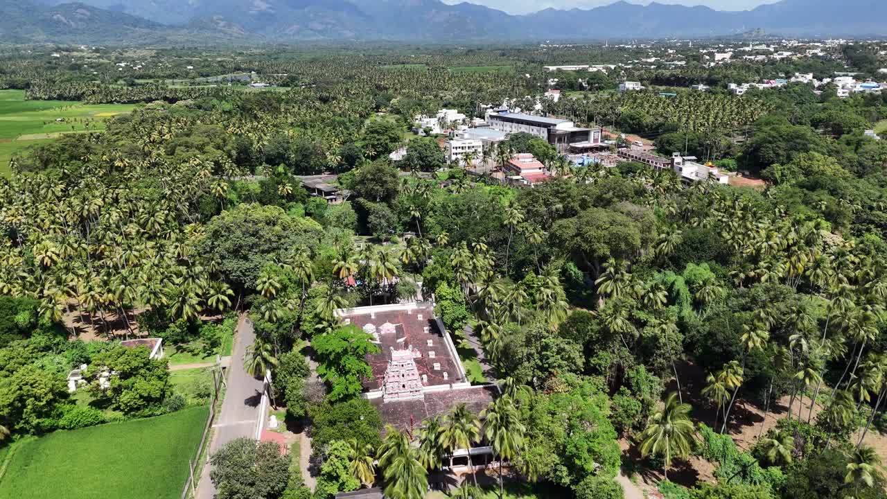 A serene aerial view captures a South Indian temple complex nestled within a landscape of lush greenery. A paved road winds through vast bright green paddy fields and coconut palm groves,