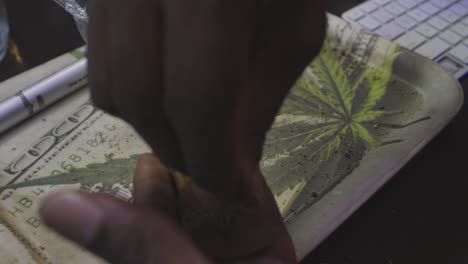 a black man sprinkles marijuana buds into a tobacco wrap to roll a blunt on a money and weed rolling tray