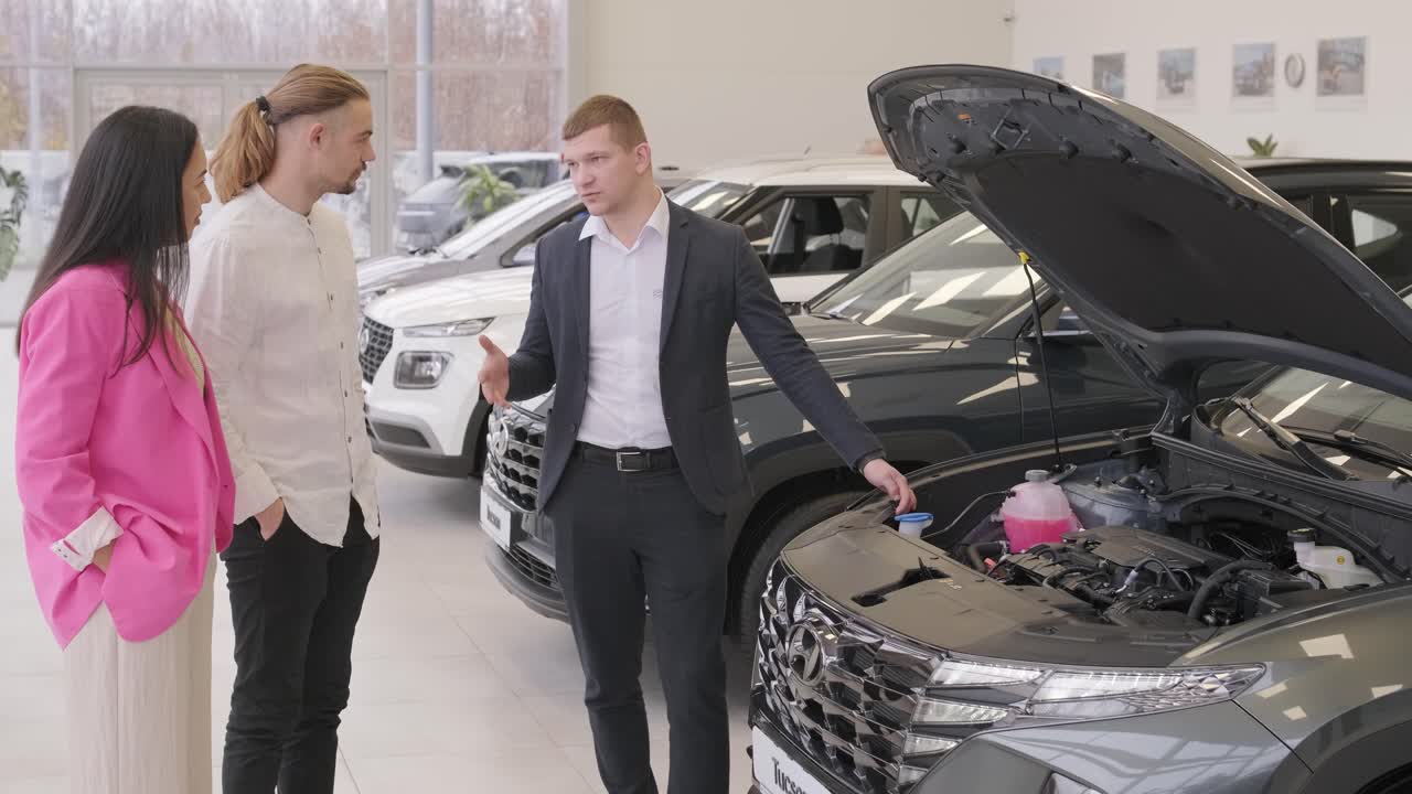 un joven vendedor de coches mostrando a una pareja de jóvenes un nuevo automóvil en el salón de concesionarios.