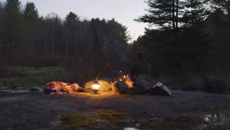 Mann-Angelt-In-Der-Abenddämmerung-Mit-Einer-Laterne-Und-Einer-Decke-Am-Lagerfeuer-In-Einem-Wald,-Umgeben-Von-Bäumen-Und-Natur