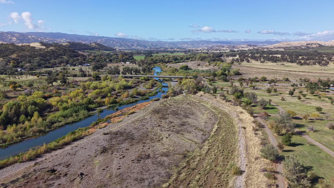 Drone performs a wide panning move above dirt roads, dry mounds and Cache Creek, revealing distant hills under a clear sky in Capay, California