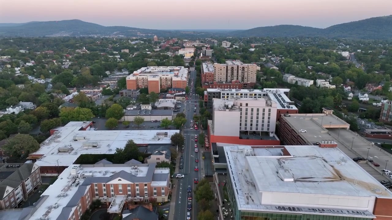 Toma aérea a vista de pájaro de la calle principal de una ciudad estadounidense al amanecer. Casas y edificios en el centro de Charlottesville, Virginia. Árboles verdes y montañas durante un día otoñal brumoso. Toma amplia.